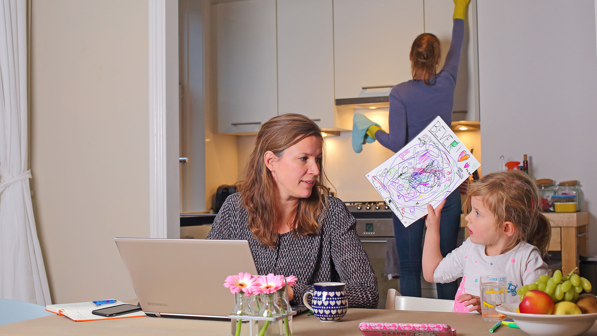 Werk en privé. Moeder aan het werk aan de keukentafel. Dochtertje is aan het kleuren.