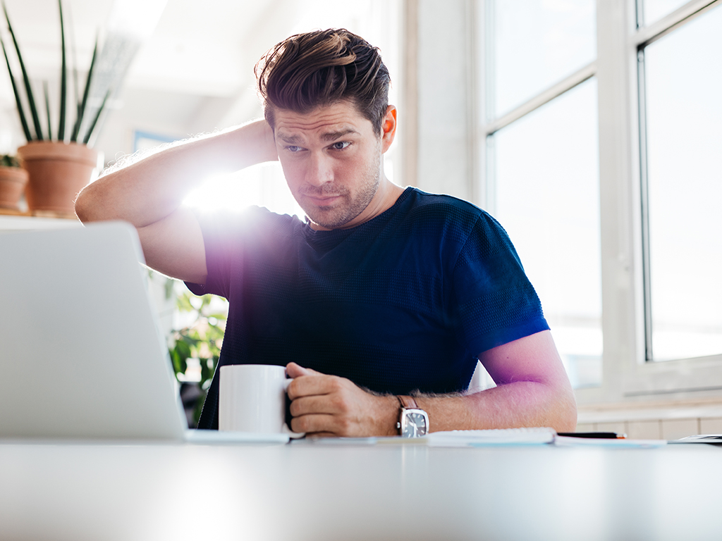 Man met beker in de hand zit in een kantoor en kijkt op een laptopscherm