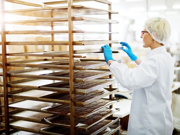 Medewerkster aan het werk in een bakkerij. Vrouw plaatst een bakplaat met koekjes in een ijzeren kar.