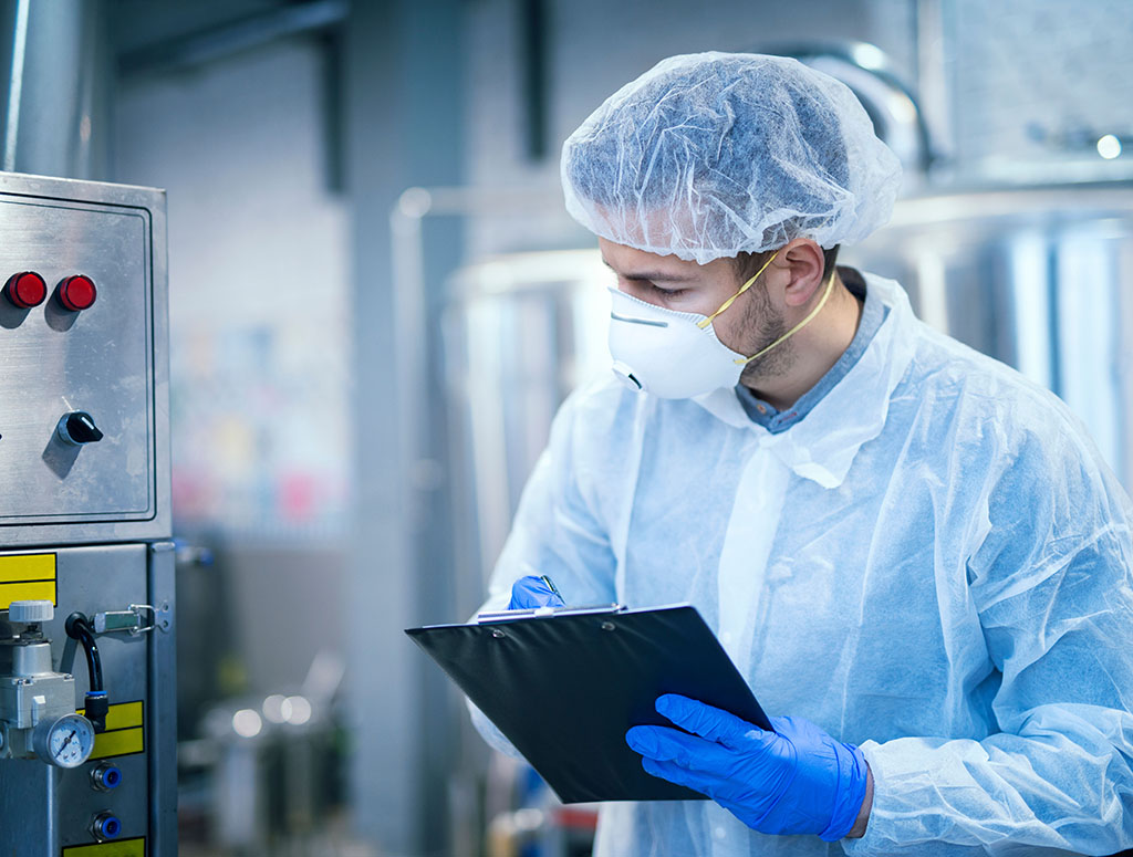 Fabrieksmedewerker in beschermende kleding controleert een apparaat en noteert dat op een klembord.