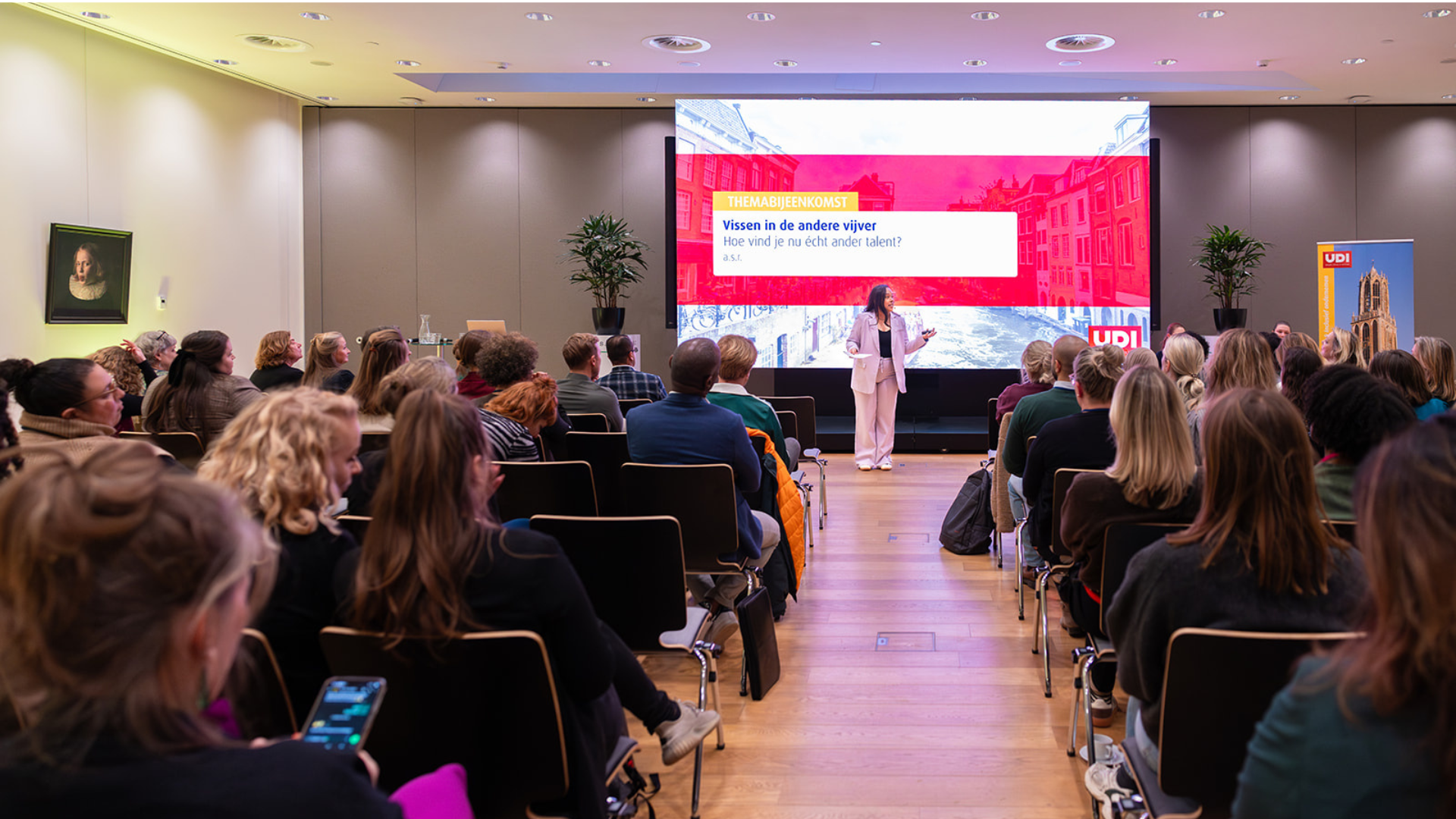 Je ziet een zaal met deelnemers en een vrouw als spreker