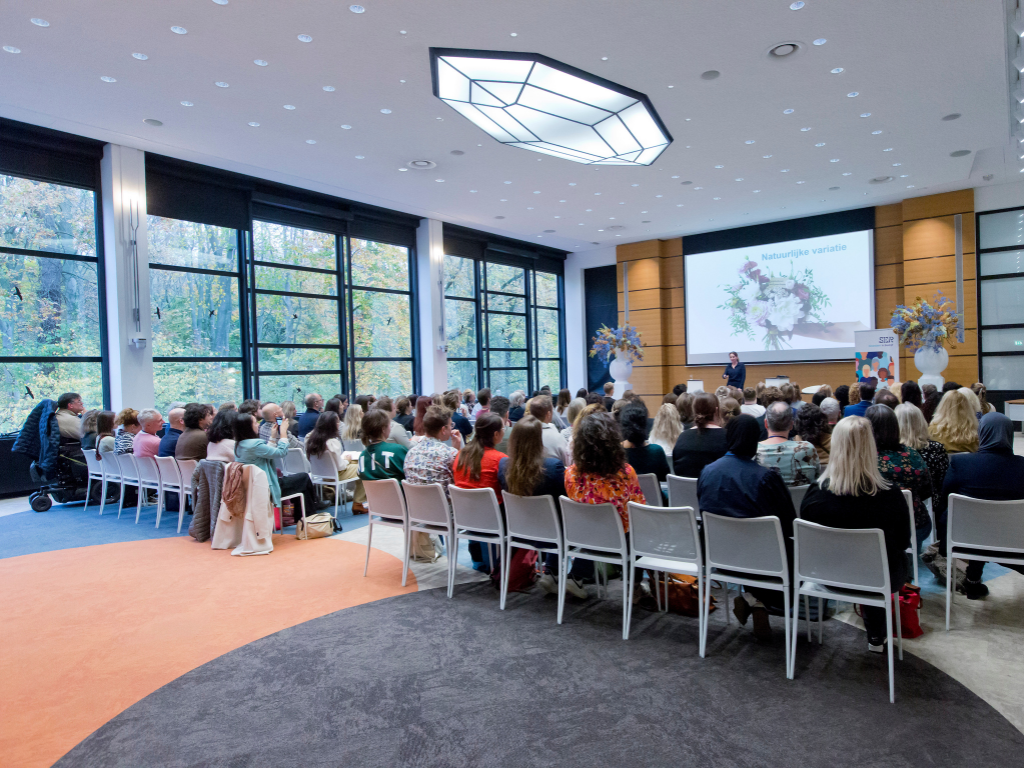 Je ziet een zaal vol mensen van achteren met een spreker voor de groep